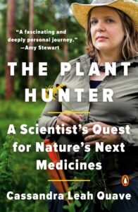 An image of a book cover titled "The Plant Hunter" in white san-serif capitals. Behind the text, a young woman in a straw hat and hiking clothes, holding a flowering plant, stares out from a backdrop of a pine forest.