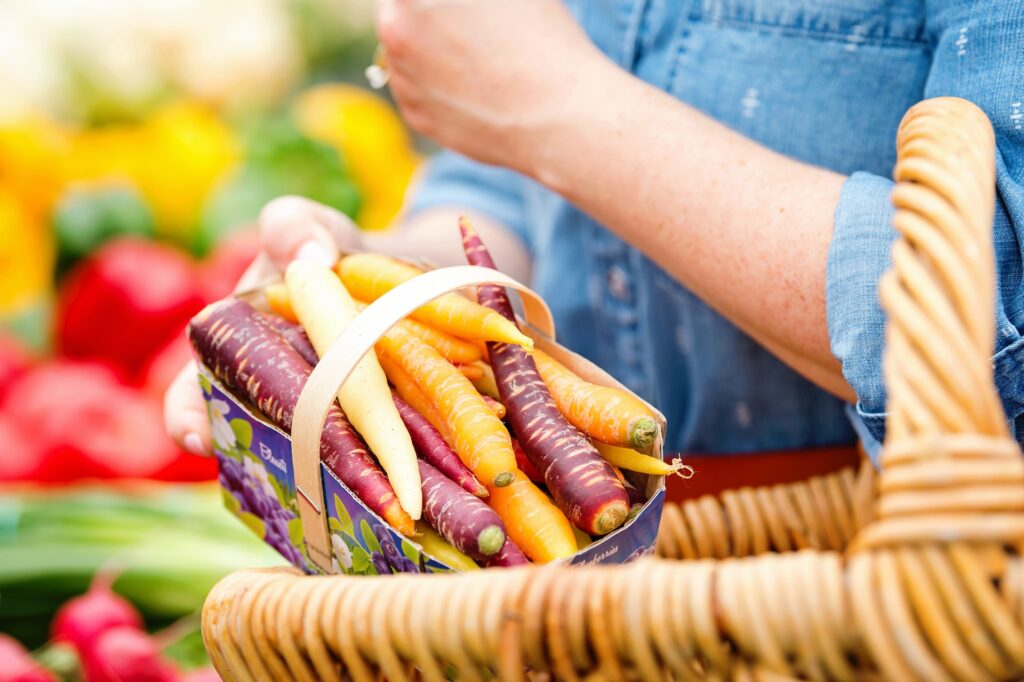 The arms and hands of a Caucasian woman wearing a blue shirt place carrots in a wicker basket.