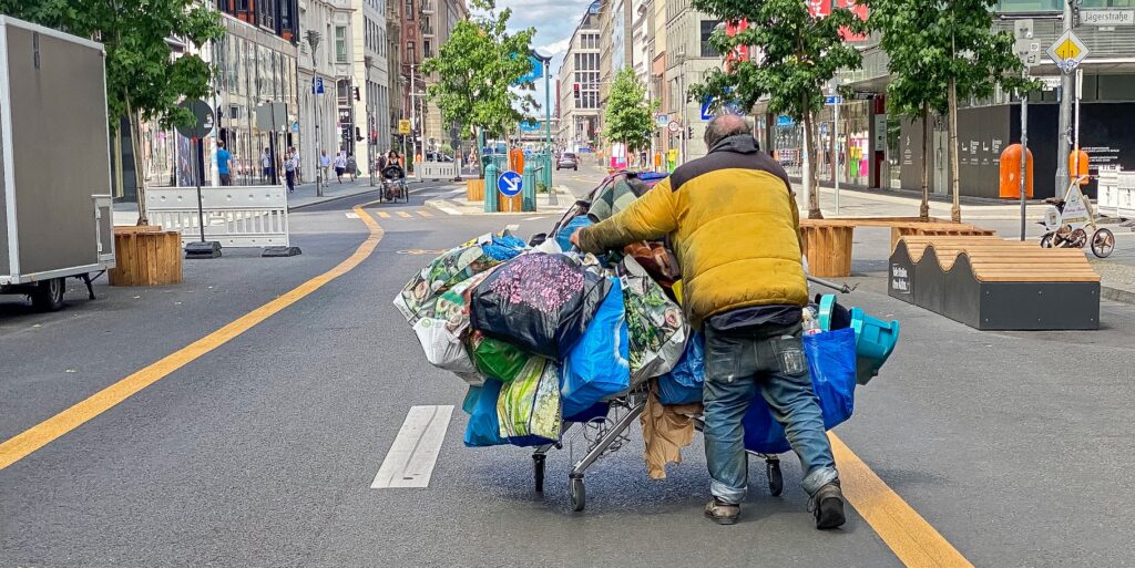 Seen from the rear, an unhoused man pushing a shopping cart laden with bags walks down a street.