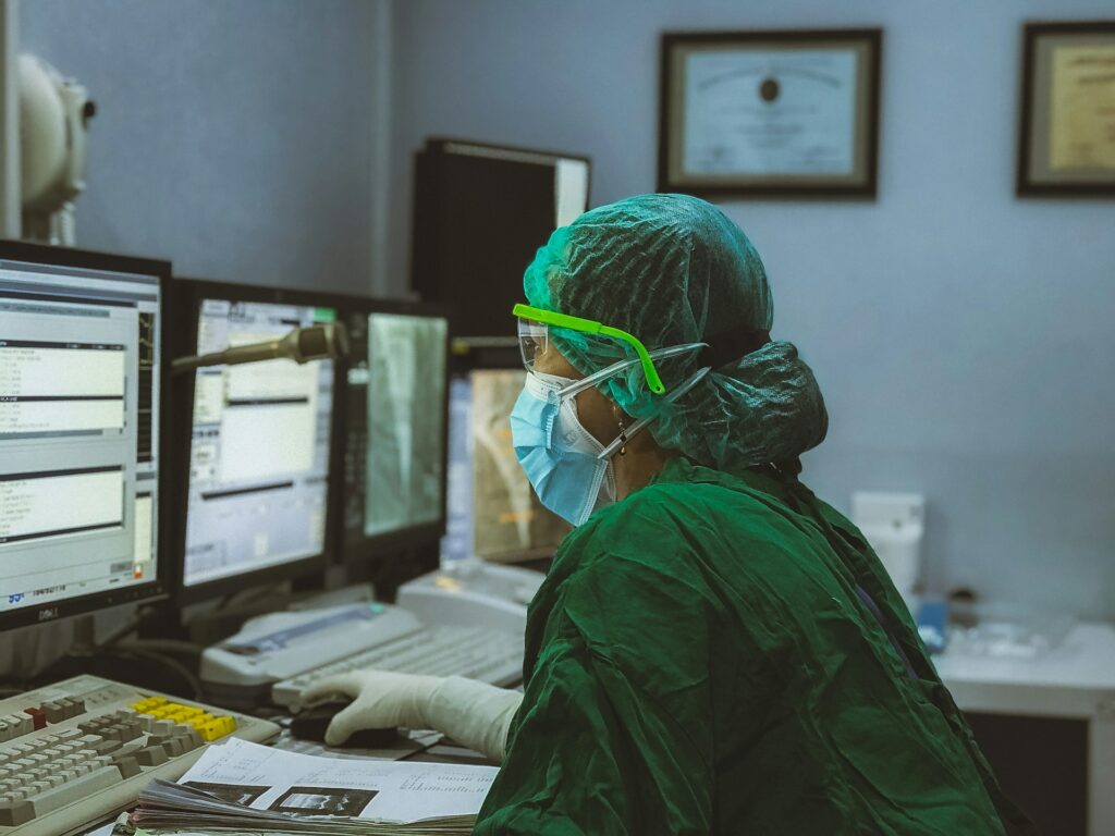In a shadowed room, a masked healthcare worker stares at computer screens.