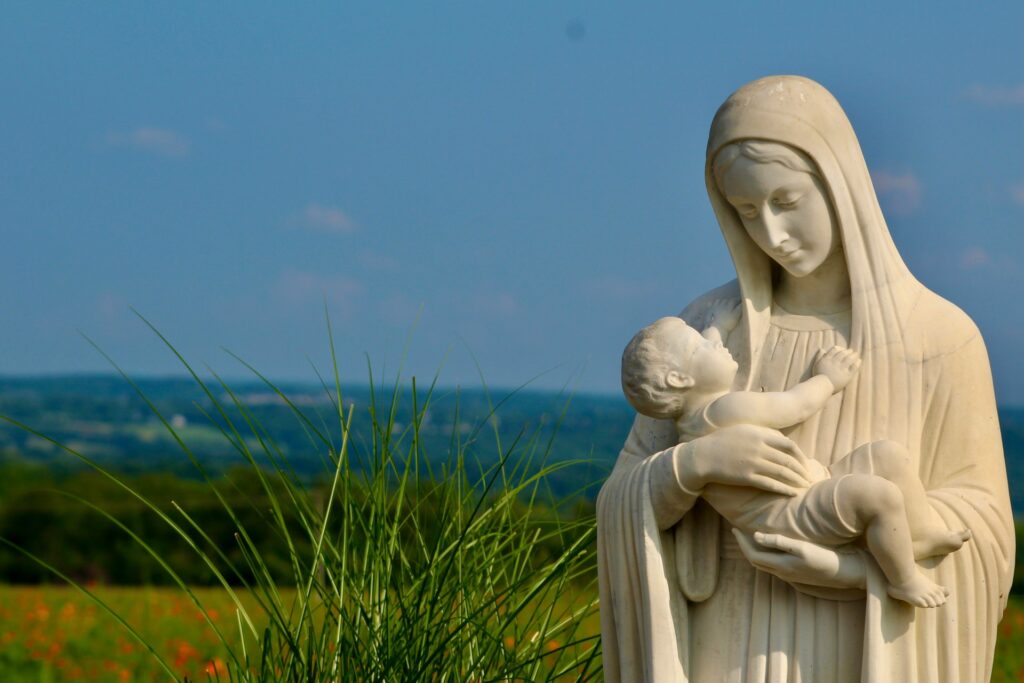 A madonna and Child statue stands against a rural backdrop of blue sky and green fields.
