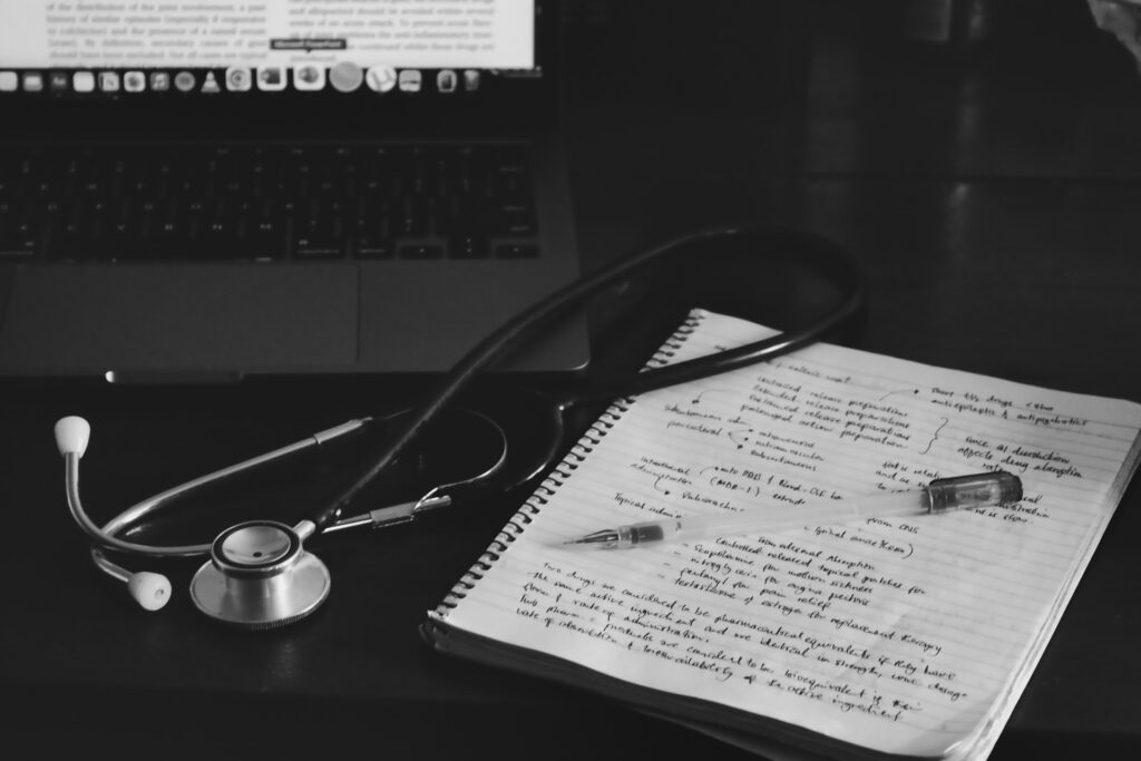 A stethoscope lies against a handwritten page of medical notes, under an out-of-focus computer screen of text.
