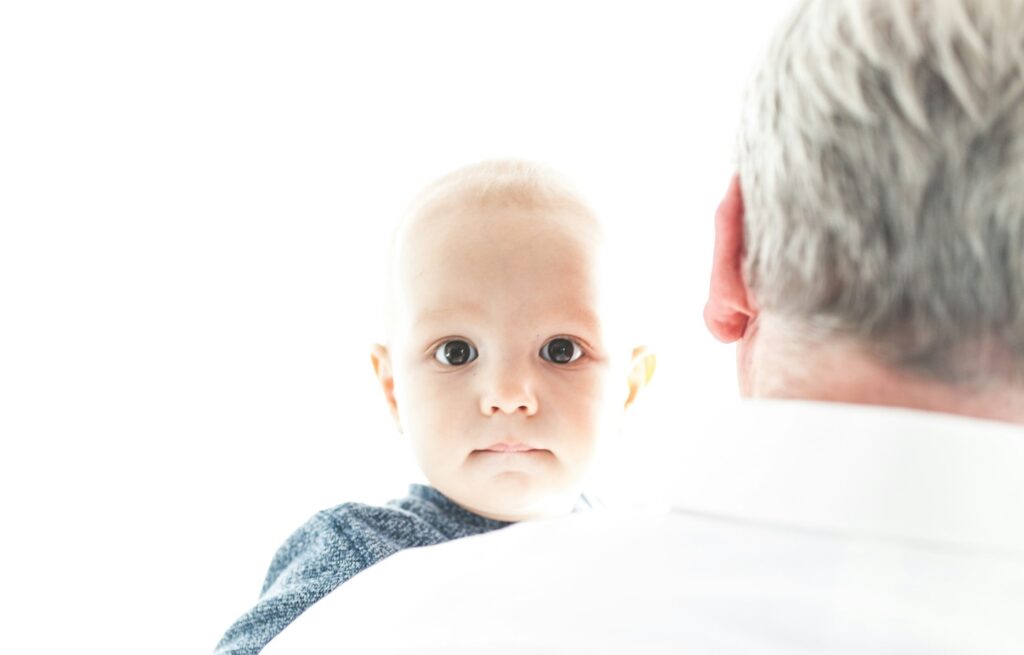 A Caucasian baby with brown eyes stares over the shoulder of an unidentified gray-haired man.