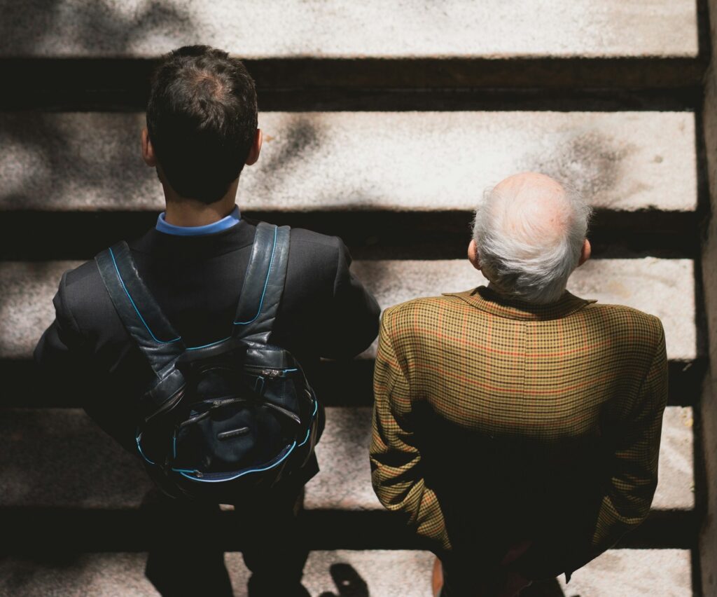 Seen from above, a dark-haired young man with a backpack and an older man in a tweed jacket walk up a set of stairs.