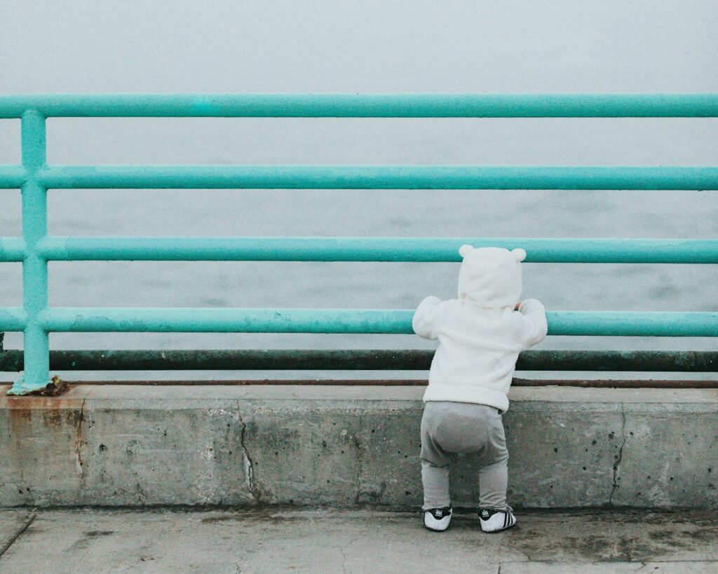 A toddler in a hoodie and baby sneakers faces away from the camera and gazes at an ocean.