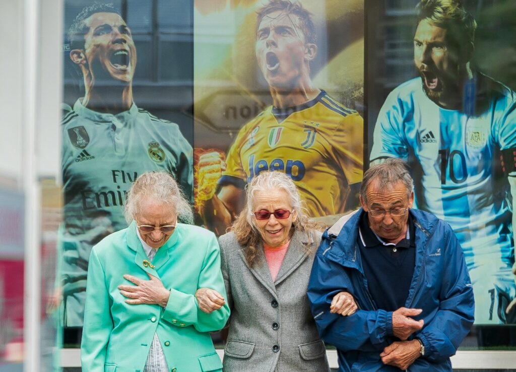 Against a backdrop of images of famous young soccer players, two older woman and an older man link arms laughing.