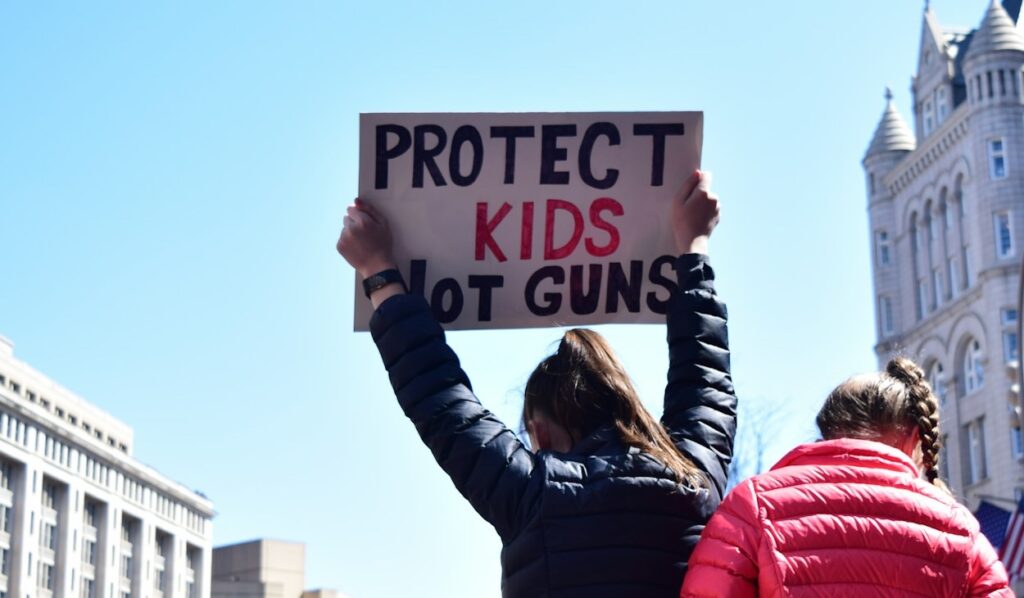 Against a blue sky, a woman with long brown hair and a black puffer jacket holds a handwritten sign that says "Protect Kids Not Guns"