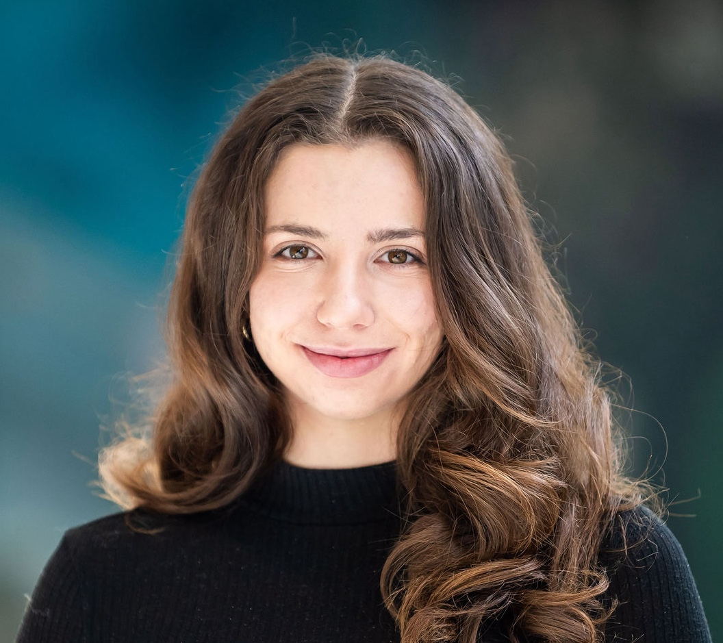 A young Caucasian woman with brown eyes and long, curling center-parted brown hair smiles against a blue background. She is wearing a long-sleeved black turtleneck.