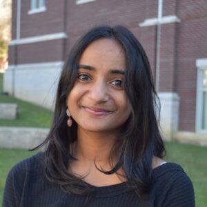 A South Asian Woman Wearing A Black Boatneck Top Smiles Against A Backdrop Of A Brick Building And Green Grass.