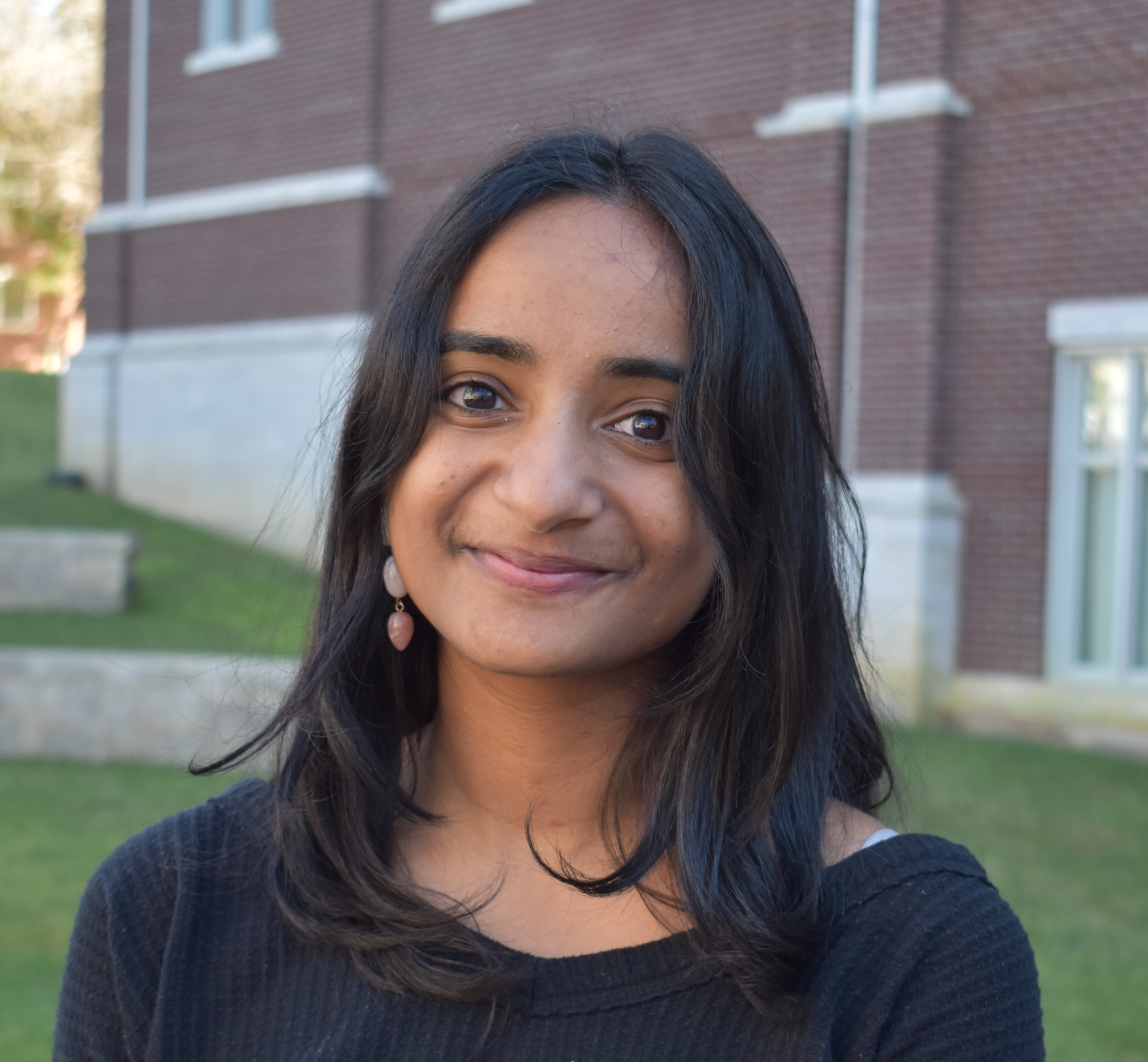 A South Asian woman wearing a black boatneck top smiles against a backdrop of a brick building and green grass.