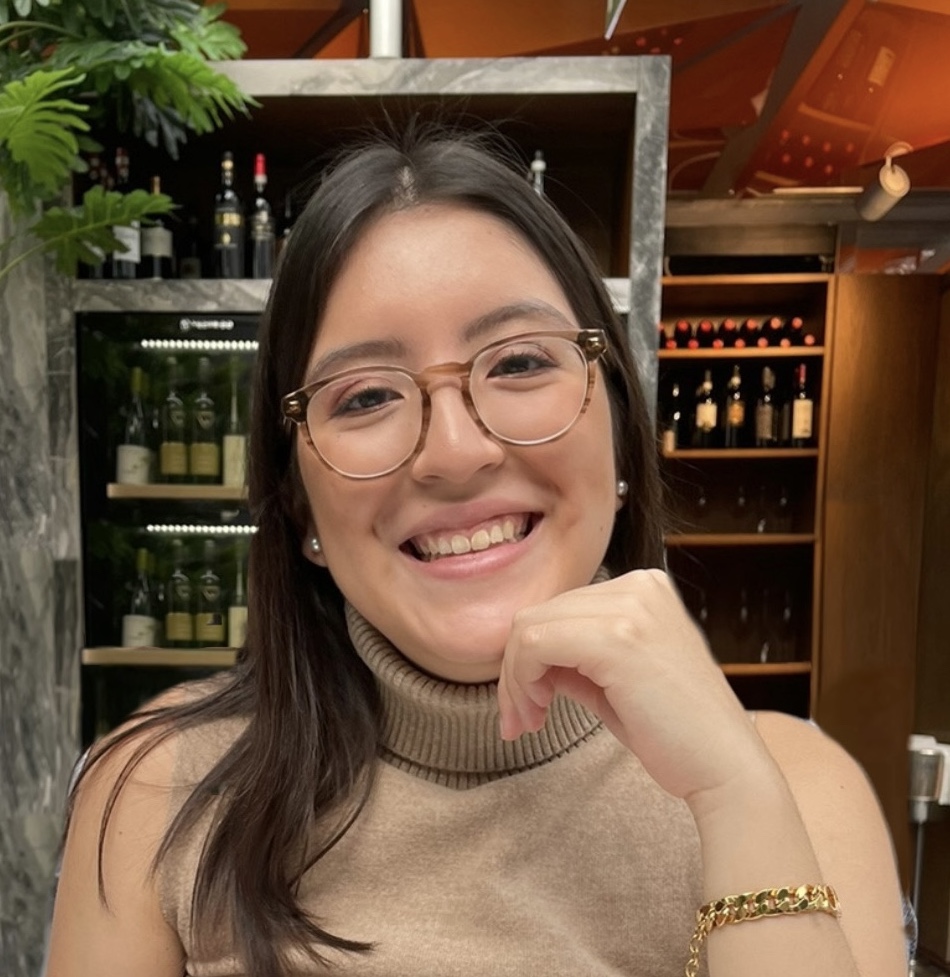 A Latina woman with dark hair and a gold chain-link bracelet smiles at the camera against a backdrop of wine bottles on shelves. She is wearing a sleeveless turtleneck that is camel-colored.