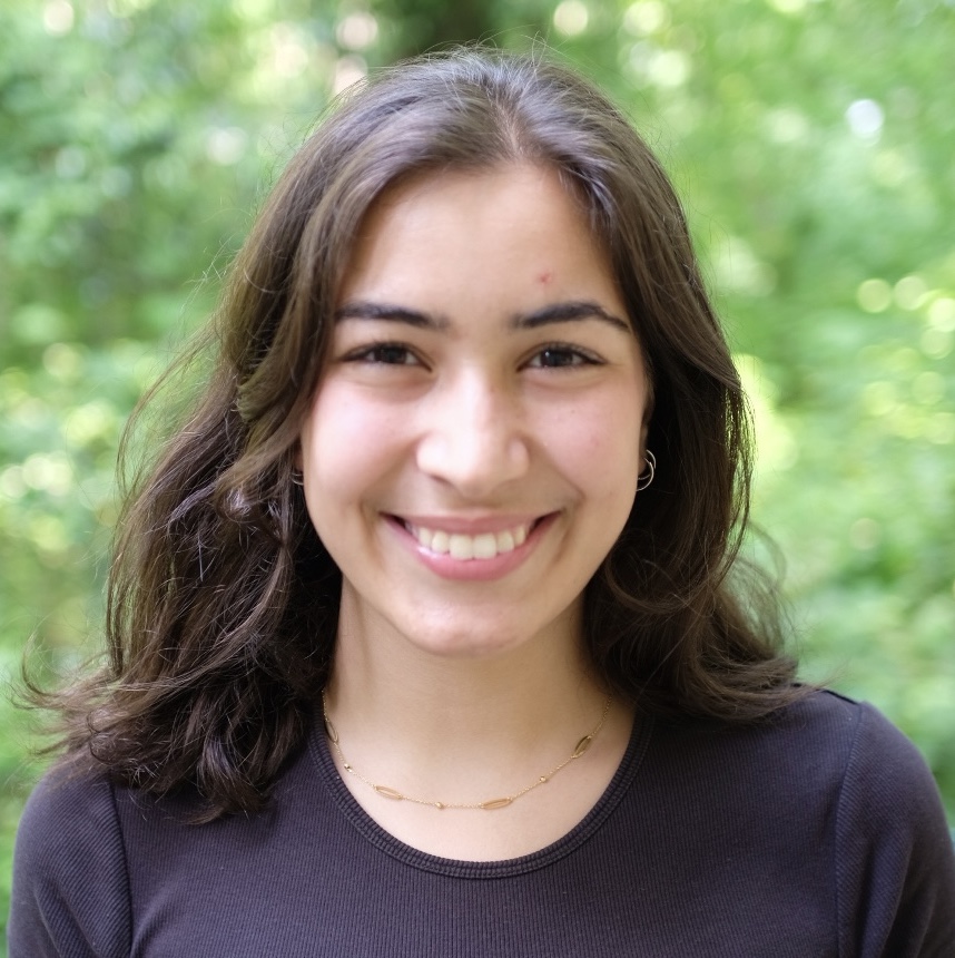 A dark-haired, dark-eyed young woman smiles against a blurred background of leaves. She is wearing a round-necked dark-blue top.