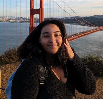 A young Black woman wearing a long-sleeved dark top smiles at the viewer. She is standing on a bluff above San Francisco's Golden Gate Bridge.