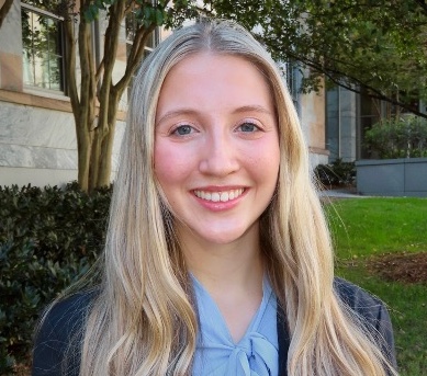 A young women with long pale blonde hair smiles at the viewer against a backdrop of trees and a stone building. She is wearing a dark blue blazer and a light blue tied blouse.